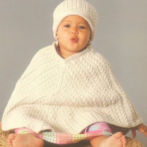 May include: A white knitted baby poncho with a cable knit pattern and a matching white knitted hat. The poncho has a v-neck and is worn by a baby sitting on a woven basket.