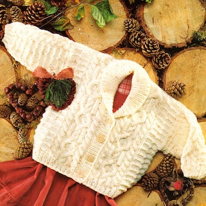 May include: A white knitted baby cardigan with a cable knit pattern and button closure. The cardigan is decorated with a red and white bow and a sprig of greenery.