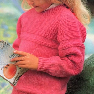 May include: A young girl wearing a pink knitted sweater with a ribbed texture. She is holding a book titled "James and the Giant Peach" by Roald Dahl.