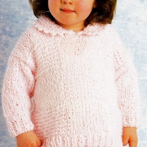 May include: A light pink knitted sweater with a ruffled collar. The sweater has long sleeves and a textured knit pattern. The child is wearing pink pants. The sweater is a soft, comfortable garment.