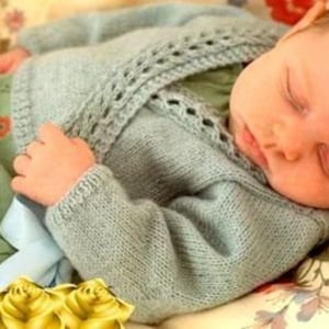 May include: A light blue knitted baby cardigan with a delicate lace trim around the neckline and sleeves. The cardigan is laid on a floral patterned fabric with yellow roses.