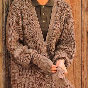 May include: A brown knitted cardigan with a cable knit pattern and a button fastening. The cardigan has pockets on the front and is worn over a black polo shirt.