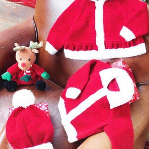 May include: A red knitted baby jacket with a white trim and hood, a matching hat with a white pom-pom, and a stuffed reindeer toy. The jacket has a ruffled hem. The items are on a brown leather surface.