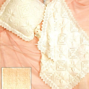 Puede incluir: Una manta de bebé de ganchillo blanco con un patrón cuadrado y una almohada a juego. La manta está sobre un fondo rosa.