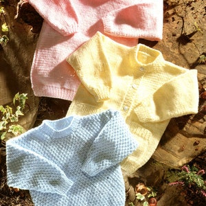 Könnte beinhalten: Drei gestrickte Baby-Pullover in Rosa, Gelb und Blau. Die Pullover liegen auf einem braunen und grünen Hintergrund.