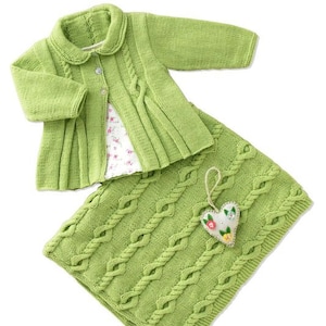 Könnte beinhalten: Ein grüner Strickcardigan mit Zopfmuster und eine passende grüne Strickdecke mit einem herzförmigen Schmuckstück.