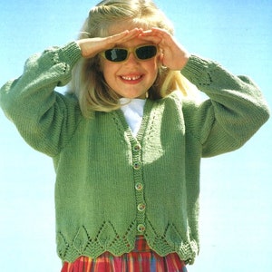 May include: A green knitted cardigan with a button-up front and a scalloped hem. The cardigan is worn by a young girl with blonde hair who is wearing sunglasses.