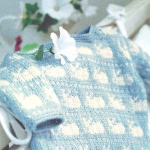 May include: A pale blue knitted jumper featuring a pattern of white shapes. The garment is displayed on a white hanger, with a white flower and green leaves resting on the shoulder. The jumper is a soft, cosy knit.