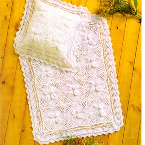 May include: A white crocheted blanket and pillow set with a floral pattern. The set is laid out on a wooden surface with a bunch of flowers.