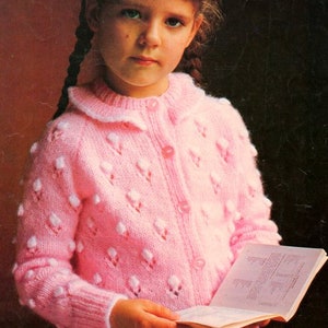 以下が含まれることがあります： 白い襟とボタン留めが付いたピンクのニットカーディガン。カーディガンは小さな白いドットの繊細なパターンがあり、長い茶色の髪の若い女の子が着用しています。