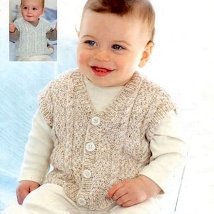 Op de afbeelding: Een close-up van een baby die een beige gebreide vest met knopen draagt. Het vest heeft een kabelbreipatroon. De baby glimlacht en kijkt naar de camera. De afbeelding is afkomstig uit een breipatronenboek met de titel "Snuggly DK 1859 Birth to 7 years".
