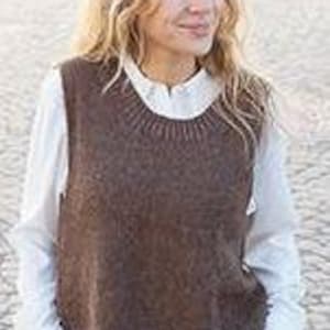 May include: A brown knitted vest with a crew neck, worn over a white collared shirt. The vest has a textured appearance and a slightly oversized fit. The model has long, wavy blonde hair.