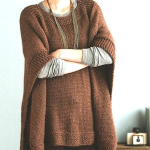 May include: A brown knitted poncho with a wide neckline and long sleeves. The poncho is worn over a grey long-sleeved shirt and brown pants. The person is wearing a black hat.