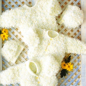May include: Three white knitted baby sweaters with a fluffy texture, laid out on a blue and white lattice background with yellow flowers.