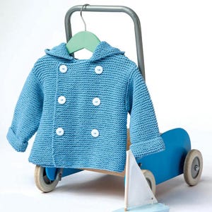 Può includere: Un cappotto per neonati in maglia blu chiaro con cappuccio e bottoni bianchi è appeso a un gancio verde. Il cappotto è esposto su un carrello giocattolo blu con una piccola barca a vela. Il cappotto è a doppio petto.
