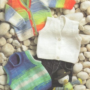 Puede incluir: Tres chalecos de punto para bebé, uno blanco, uno a rayas azules y verdes, y uno a rayas naranjas, blancas y azules. Los chalecos están dispuestos sobre un fondo de pequeñas piedras grises.