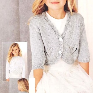 May include: A young girl wearing a light gray knitted cardigan with a white button closure. The cardigan has a delicate lace pattern and is cropped at the waist. The girl is also wearing a white lace skirt.