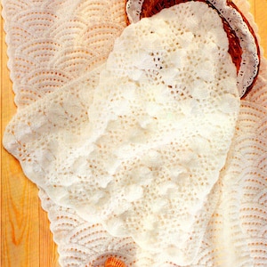 Op de afbeelding: Een wit gehaakt babydekentje met een delicate kantversiering. Het dekentje is gevouwen en gedrapeerd over een houten oppervlak met schelpen eromheen.