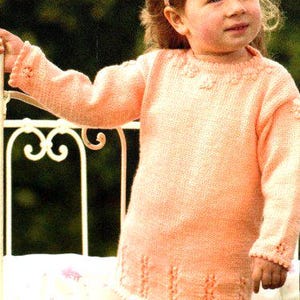May include: A young girl wearing a peach-coloured knitted jumper with a white headband. The jumper has a delicate floral pattern around the neckline and a textured design at the hem.