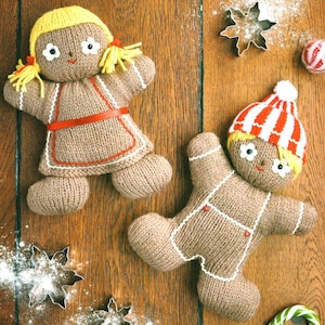 May include: Two knitted gingerbread people dolls with yellow yarn hair and smiling faces. One doll wears a red apron, and the other wears a red and white striped hat. The dolls are on a wooden surface with snowflake cookie cutters and candy canes.