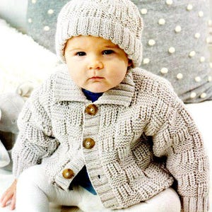 Può includere: Un bambino con un cappello di lana grigio chiaro con un pompon e un cardigan abbinato. Il cardigan ha un colletto e tre bottoni marroni. Il bambino indossa anche leggings bianchi.