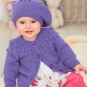 Könnte beinhalten: Eine lila Strickjacke mit Knöpfen und einer passenden lila Strickmütze. Die Strickjacke hat ein strukturiertes Muster mit kleinen, erhabenen Punkten.