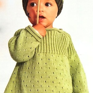 May include: A green knitted sweater with a textured pattern. The sweater has a round neckline and long sleeves. The sweater is being worn by a child who is wearing a striped hat with a pom-pom.