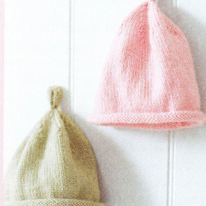 May include: Two knitted baby hats. One is light pink, the other is beige. Both hats have a rolled brim and a small loop at the top. The hats are displayed against a white background.