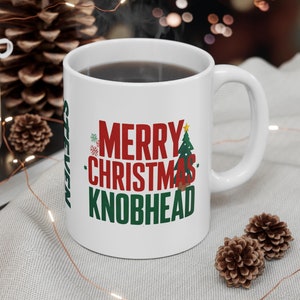 May include: White ceramic mug with the text "MERRY CHRISTMAS KNOBHEAD" in red and green. The mug also has the name "STEVEN" printed vertically in green. A Christmas tree and snowflakes are included in the design. The mug is filled with a dark liquid.
