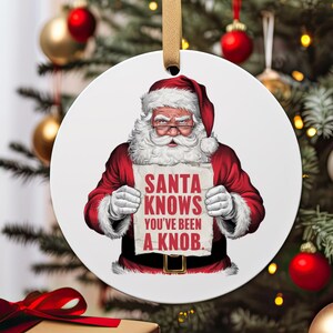 May include: Round white Christmas bauble featuring a cartoon illustration of Father Christmas holding a sign that reads "Santa Knows You've Been A Knob."
