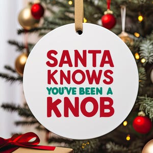May include: White round Christmas bauble with the text "Santa Knows You've Been A Knob" in red and green lettering.