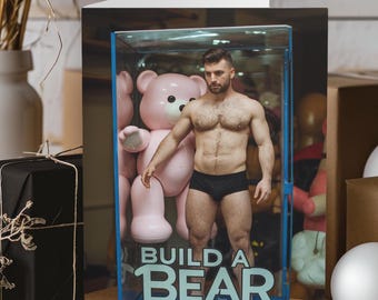 Puede incluir: Una tarjeta de felicitación con un hombre de pie en una vitrina de vidrio con un gran oso de peluche rosa. El texto "BUILD A BEAR" está impreso en la parte inferior de la vitrina.