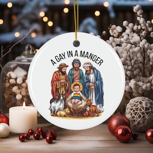May include: A white ceramic Christmas bauble featuring a colourful illustration of a nativity scene. The text "A Gay in a Manger" is printed in black at the top of the bauble.