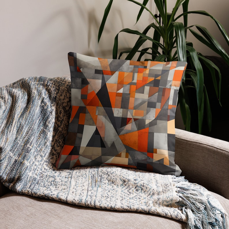 May include: A throw pillow with a geometric pattern in shades of orange, gray, and black. The pillow is on a couch with a gray and white throw blanket.