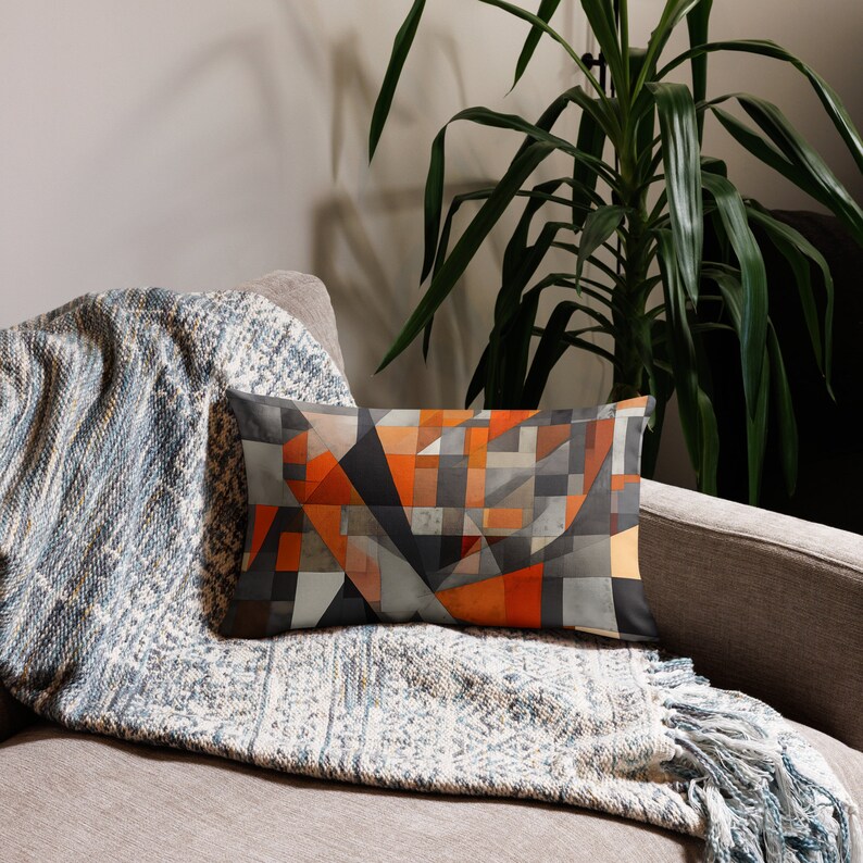 May include: A rectangular throw pillow with a geometric pattern in shades of orange, gray, and black. The pillow is on a couch with a blue and white throw blanket.