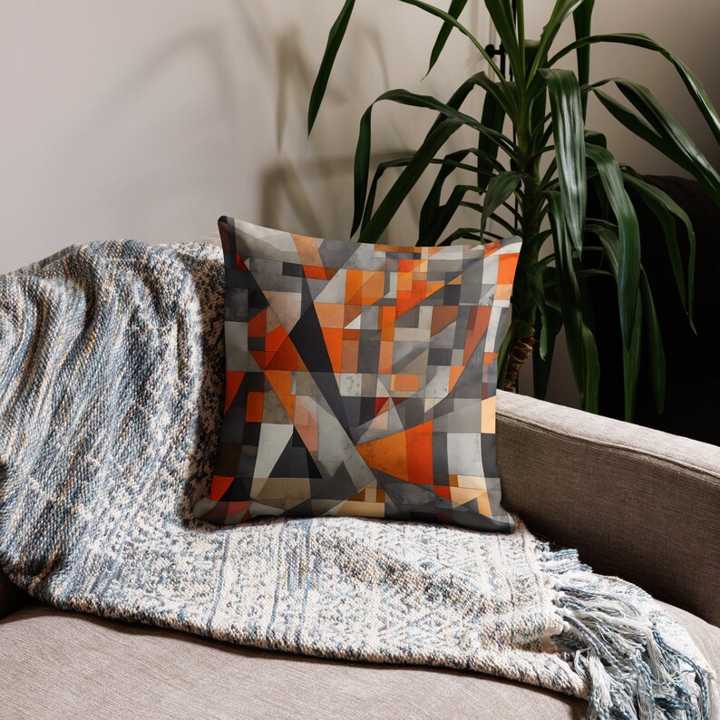 May include: A throw pillow with a geometric pattern in shades of orange, gray, and black. The pillow is on a couch with a gray and white throw blanket.