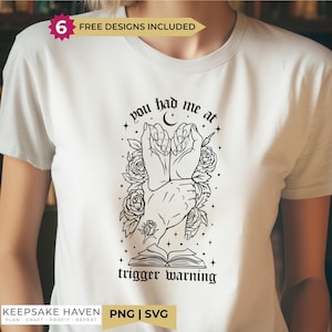 Può includere: Una t-shirt bianca con una stampa grafica nera di due mani unite, circondate da rose, una luna crescente e un libro aperto. Il testo recita "you had me at trigger warning". Un badge in alto riporta la scritta "6 FREE DESIGNS INCLUDED".