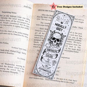 Puede incluir: Un marcapáginas de papel rectangular con una ilustración en blanco y negro de un cráneo humano rodeado de mariposas, rosas y un libro abierto. El texto dice MORALLY GREY IS MY FAVORITE COLOR. Está colocado dentro de un libro abierto.