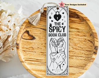 May include: A rectangular white bookmark with a black line art design featuring a heart-shaped padlock, flames, and hands with the text "THE SPICY BOOK CLUB" and "good girl". A beige tassel is attached to the top hole of the bookmark.
