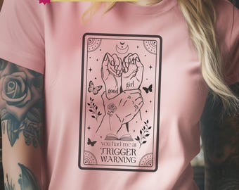 May include: A light pink t-shirt featuring a black graphic print of a tarot card design. The illustration depicts two hands holding each other with the text good girl, a rose, butterflies, and the text you had me at TRIGGER WARNING.