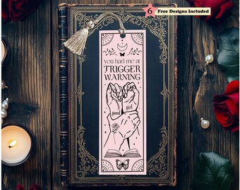 May include: A rectangular light pink bookmark featuring a black line art illustration of two hands holding each other, with the text "you had me at TRIGGER WARNING" and "good girl" written on the hands, topped with a metallic gold tassel.