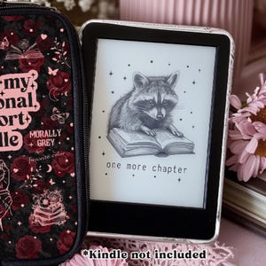 May include: A black e-reader case featuring a pattern of dark red roses, butterflies, and books, with text reading "my personal library" and "morally grey". Beside it, an e-reader displays a black and white illustration of a raccoon reading a book.