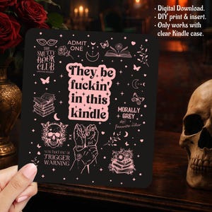 以下が含まれることがあります： 黒い長方形のKindle用インサート。ドクロ、本、蝶、月、星などのライトピンクのグラフィックイラストが描かれています。テキストには「The Smutty Book Club」「Admit One」「They be fuckin' in this kindle」「Morally Grey」「Trigger Warning」と記されています。