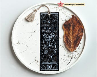 May include: A rectangular black bookmark featuring white line art of hands, a rose, an open book, and a butterfly. The text reads "you had me at TRIGGER WARNING" and "good girl". A beige tassel is attached to the top hole of the bookmark.