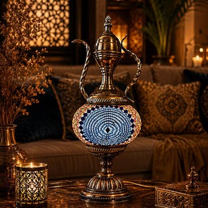 Puede incluir: Una lámpara de mesa decorativa con forma de jarra tradicional, que presenta un marco de metal en tono latón y una pantalla central en forma de globo cubierta con un mosaico de pequeñas piezas de vidrio azul, blanco y naranja.