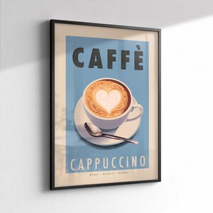 Op de afbeelding: Ingelijste kunstprint met een cappuccino met hartvormige latte art. De poster heeft een lichtblauwe achtergrond met het woord "CAFF&Egrave;" bovenaan en "CAPPUCCINO" onderaan. Een witte kop en schotel met een lepel staan in het midden.