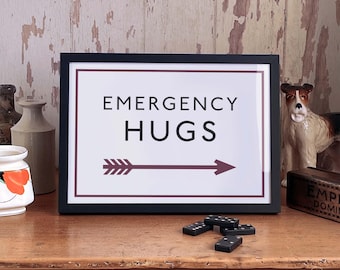 May include: A framed print with a white background and black text that reads "Emergency Hugs" with a maroon arrow pointing to the right. The print is in a black frame and is sitting on a wooden surface.