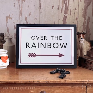 May include: A black framed print with a white background and the text "Over the Rainbow" in black letters. A maroon arrow points to the right. The print is on a wooden surface with dominoes and a ceramic dog figurine.