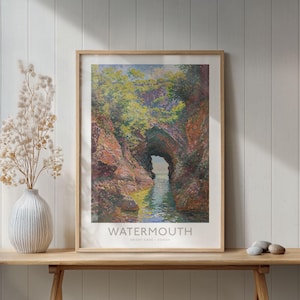 Op de afbeelding: Een ingelijste print van Watermouth, Devon, met een landschapsschilderij van een kustgrot. Het kunstwerk heeft een lichte houten lijst en wordt tentoongesteld op een houten tafel. De print bevat de tekst "WATERMOUTH" en "Hele Bay - Devon".