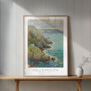 May include: Framed art print of Hell's Mouth, a coastal landscape. The artwork depicts a rocky coastline with green vegetation and turquoise water. The print is in a light wood frame and is displayed on a wooden table.